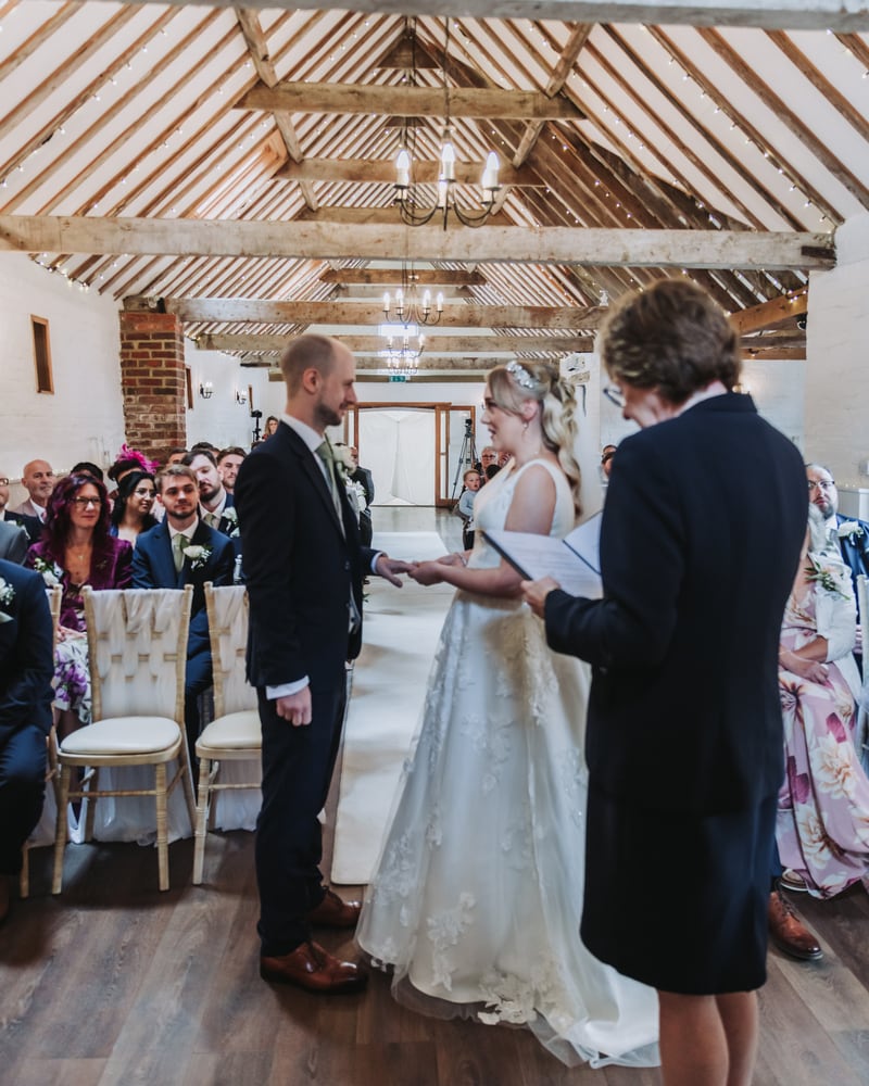 A couple saying their vows in the barn beneath oak roof trusses and chandeliers, guests seated either side
