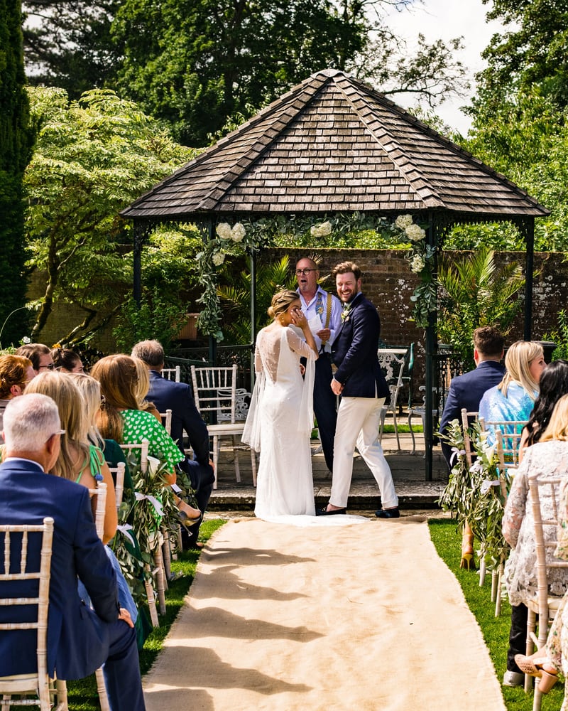 A couple exchanging vows beneath the slate-roofed gazebo, guests seated either side of a hessian aisle
