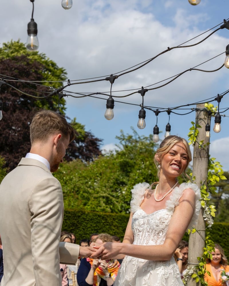 A laughing bride and groom holding hands beneath festoon lights strung from the maypole