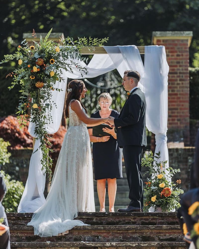 A bride and groom holding hands with their celebrant beneath a draped, sunflower-dressed arch at the top of the brick steps
