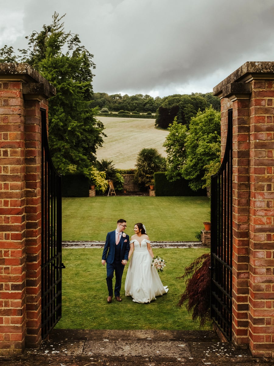 A bride and groom walking hand in hand between the brick gate piers, the lawn and Kent hills behind