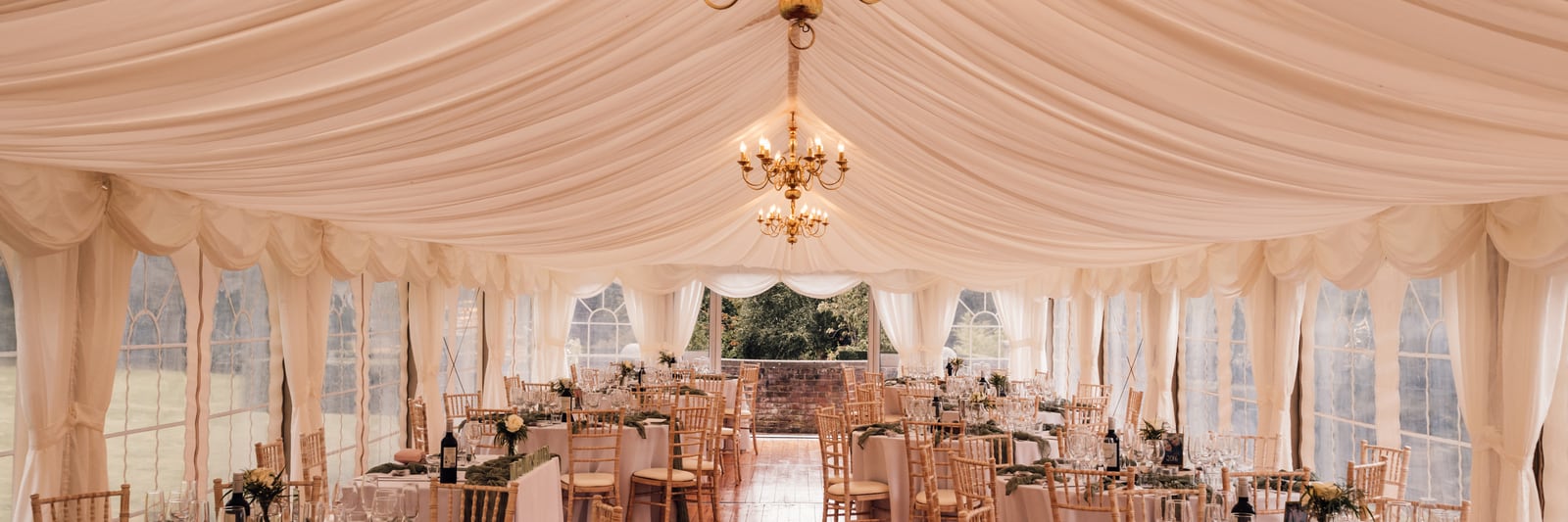 The marquee laid for the wedding breakfast, warm light from twin chandeliers over white linen and chiavari chairs